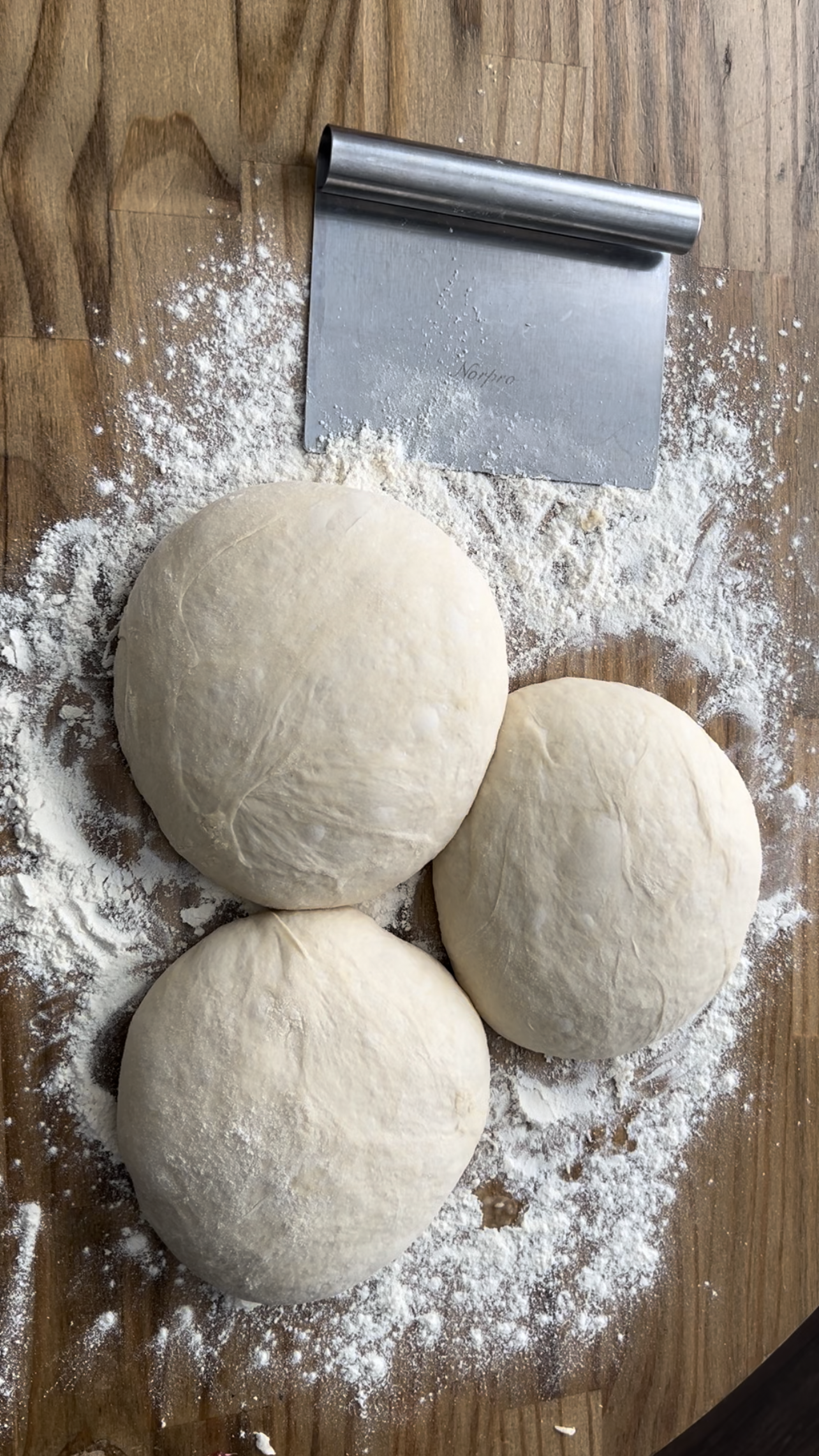 The perfect pizza dough in 3 sections on a floured surface with a bench scraper next to them #perfectpizzadough #pizza #pizzacrust #neopolitanstylepizza